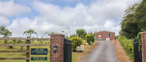 Una entrada rural con una cancela, un cartel que dice 'Car Park' y un edificio con el letrero 'Open Times'.