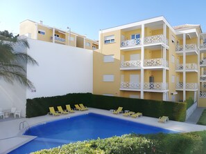 Une piscine avec des chaises longues, un mur blanc et un bâtiment jaune doté de balcons.