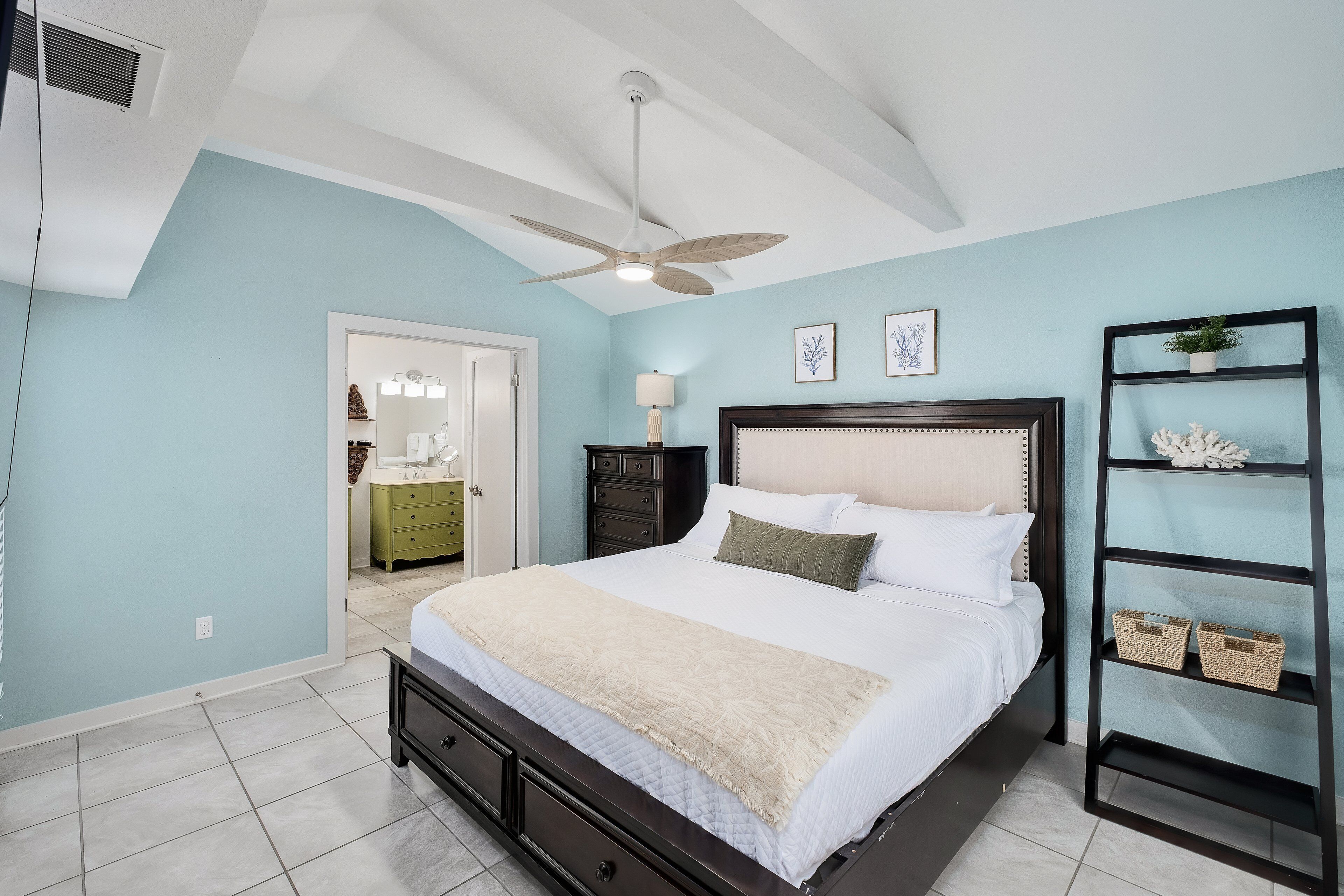 A bedroom with a bed, a ceiling fan, a nightstand, a dresser, a shelf with baskets, and a door leading to another room.