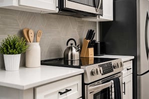 A modern kitchen with a stainless steel stove, microwave, and refrigerator, wooden utensils, and a potted plant. - Boulevard Stays Quiet Modern 1BR with Pickleball, Pool, Parking & Gym (Chandler)