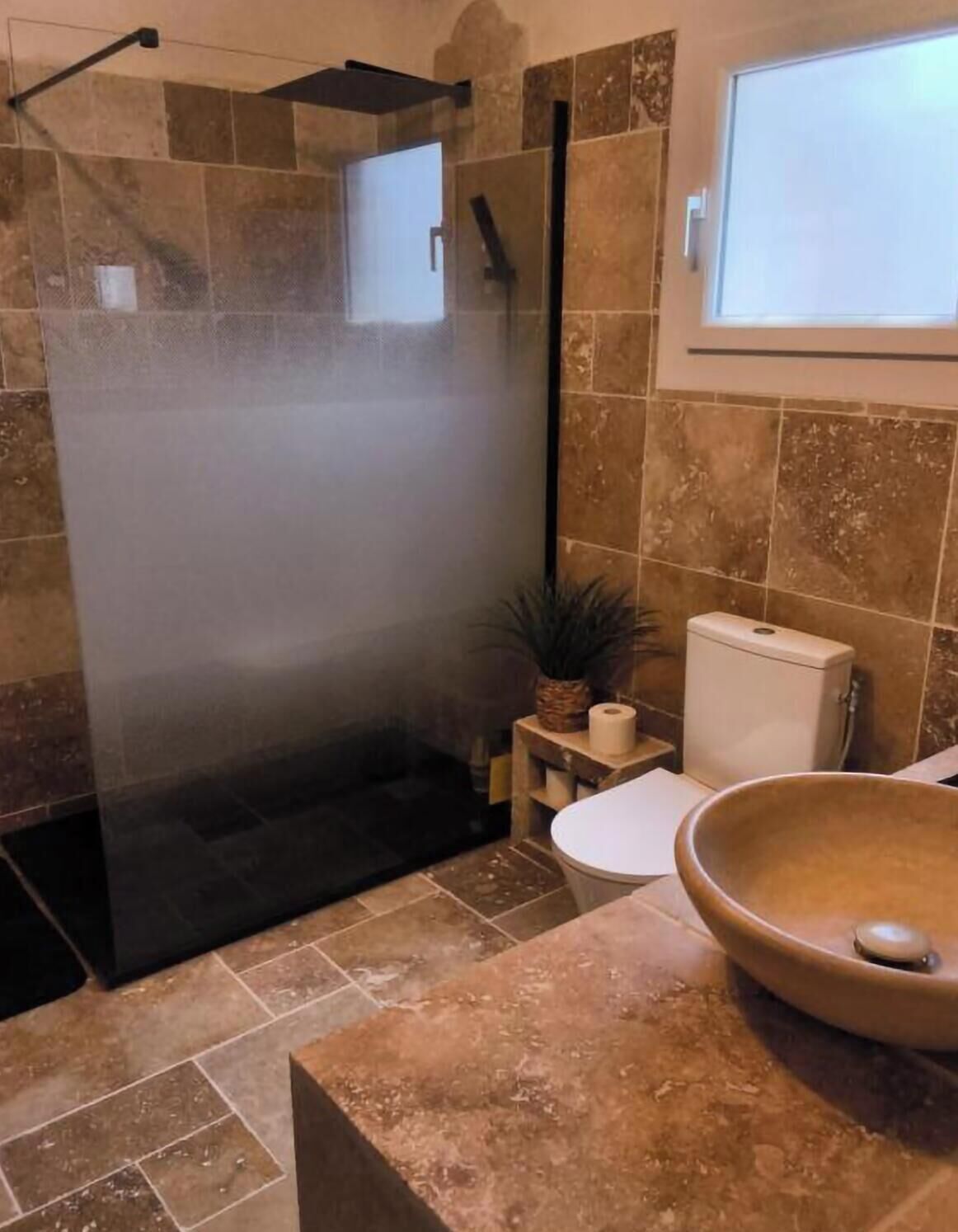A bathroom with a shower enclosure, a toilet, a sink with a bowl, and a window.