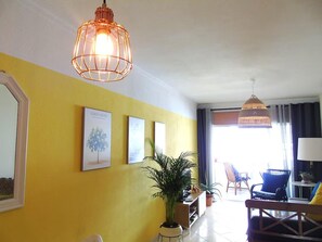 A room with a yellow wall, a hanging light fixture, framed artwork, a potted plant, and a seating area with a chair and a table. - 2 bed - Sleeps 4 - Balcony - City Centre (Faro)