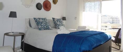 A bedroom with a bed, pillows, a bedside table, a lamp, a wall-mounted air conditioner, and a window with a view.