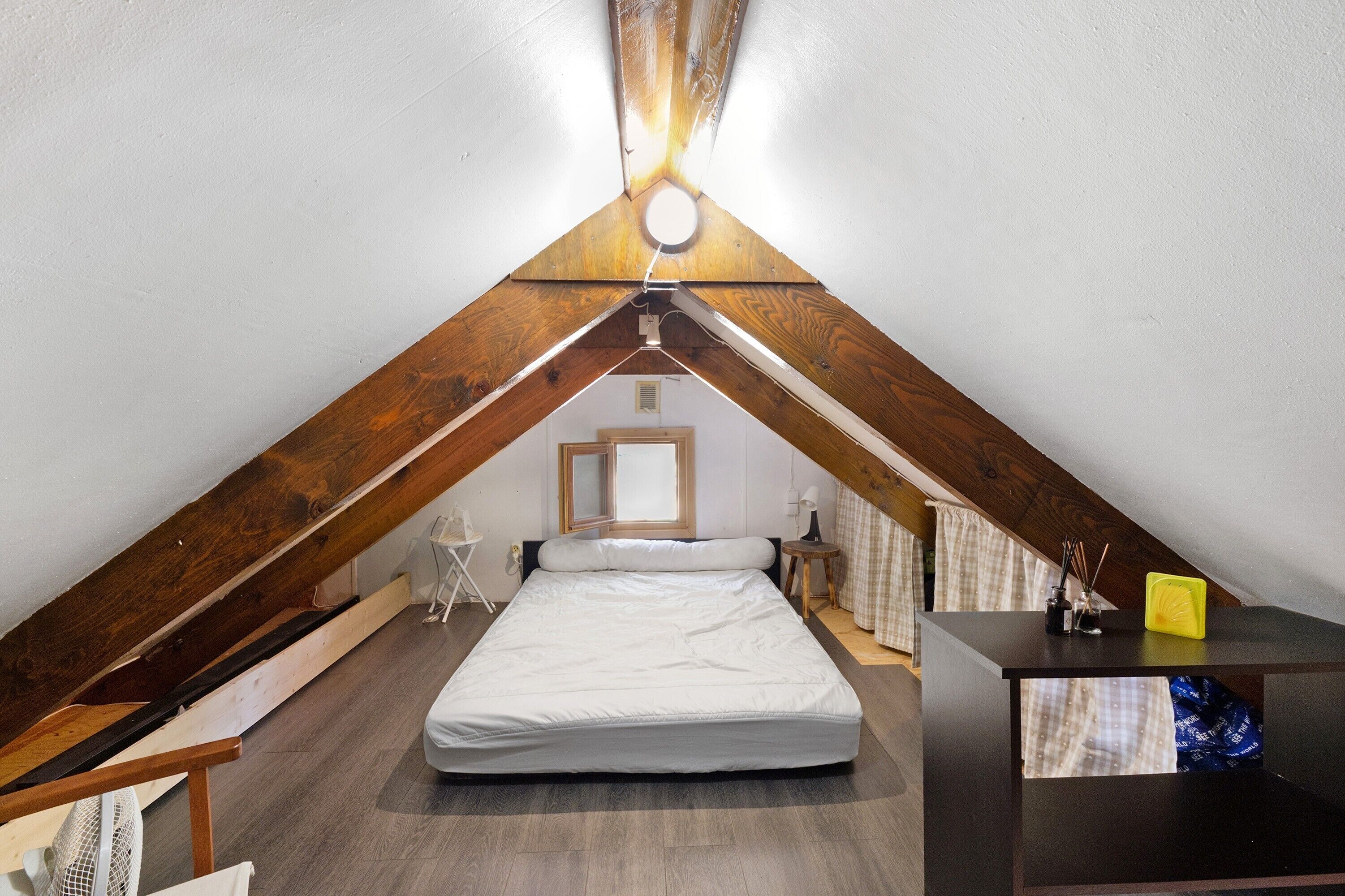 Une petite chambre mansardée de forme triangulaire, avec un lit simple, un bureau et un luminaire suspendu.