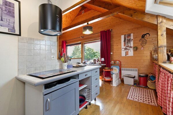 A kitchen with a tiled backsplash, wooden ceiling beams, a window with red curtains, and a small refrigerator.