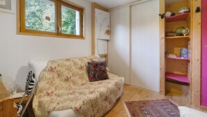 Ett mysigt rum med en soffa med blommönster, en liten matta, en trähylla med böcker och dekorativa föremål samt ett fönster med utsikt över grönskan.