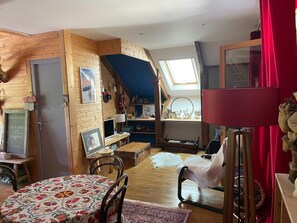 Une piĂšce chaleureuse avec un lit en mezzanine, des murs en bois, une lucarne, un rideau rouge et une petite table avec une chaise.