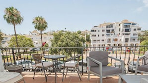 A balcony with chairs and a table, overlooking a street with parked cars and buildings.