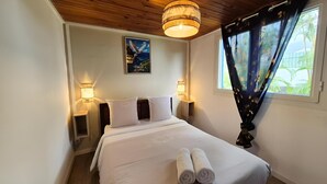 A neatly made bed with white linens and two rolled towels, a wooden ceiling, a wall-mounted painting, and a window with curtains.