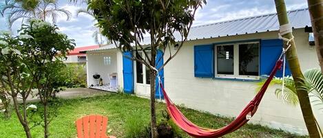 A small building with a white exterior and blue shutters, surrounded by greenery and a hammock.