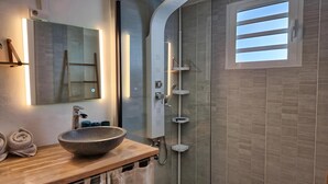 A modern bathroom with a large mirror, a wooden vanity, a stone basin, a glass-enclosed shower, and a view of the sky through a window.