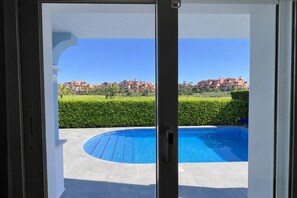 On aperçoit, à travers une porte vitrée, une piscine, un ciel dégagé et, au loin, des immeubles d’habitation.
