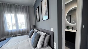 Une chambre moderne avec un grand lit, une table de chevet, une lampe et une salle de bain équipée d’un miroir et d’un lavabo.