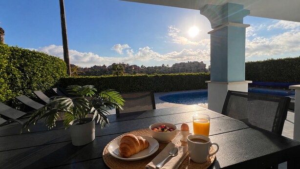 En uteservering med utsikt över en swimmingpool och byggnader, ett croissant, en skål med frukt och ett glas apelsinjuice.