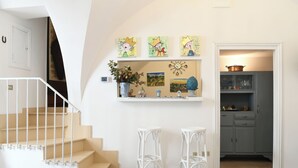 Uma escada leva a um patamar com corrimão branco, uma prateleira com obras de arte e um vaso, e uma área de cozinha com armários e uma panela.