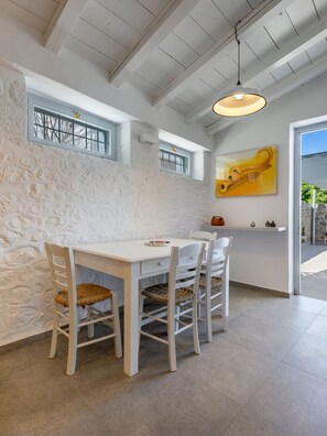 A dining area with a white table and chairs, a hanging light fixture, a stone wall, and a window with a view. - Hydrelion Beachfront Suite, Divine Sea Views, Furnished Terrace, Hydra (Hydra)