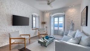 A modern living room with a stone-textured wall, a flat-screen TV, a ceiling fan, a sofa, a coffee table, and a view of the sea through large windows. - Beachfront Villa Ondyrelis, Divine Sea Views, Furnished Terrace, Hydra (Hydra)