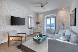 A modern living room with a stone-textured wall, a flat-screen TV, a ceiling fan, a sofa, a coffee table, and a view of the sea through large windows. - Beachfront Villa Ondyrelis, Divine Sea Views, Furnished Terrace, Hydra (Hydra)