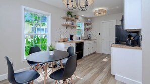 Een moderne keuken met witte kasten, een eethoek met een ronde tafel en stoelen, en een groot raam met uitzicht op groen.