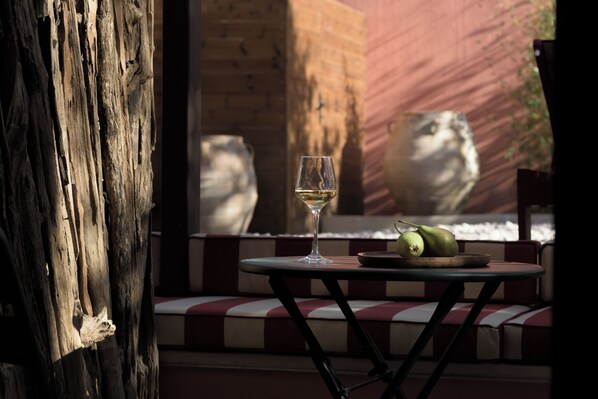 A table with a wine glass and fruit, a wooden bench, and a stone sculpture in the background.