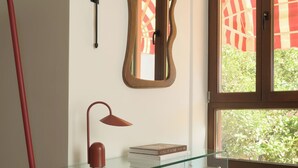 Una habitación con un escritorio de vidrio, una lámpara roja, un espejo y una ventana con vistas a la vegetación.