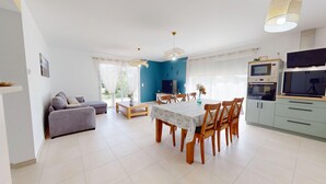 A modern living room with a sofa, a dining table with chairs, a TV, and a kitchen area with cabinets and appliances. - Serenity – Family home with a garden and Wi-Fi in (Saint-Jean-de-Monts)