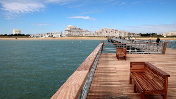 A wooden pier with benches extending over calm water, leading to a beach with buildings in the background. - Serenity – Family home with a garden and Wi-Fi in (Saint-Jean-de-Monts)