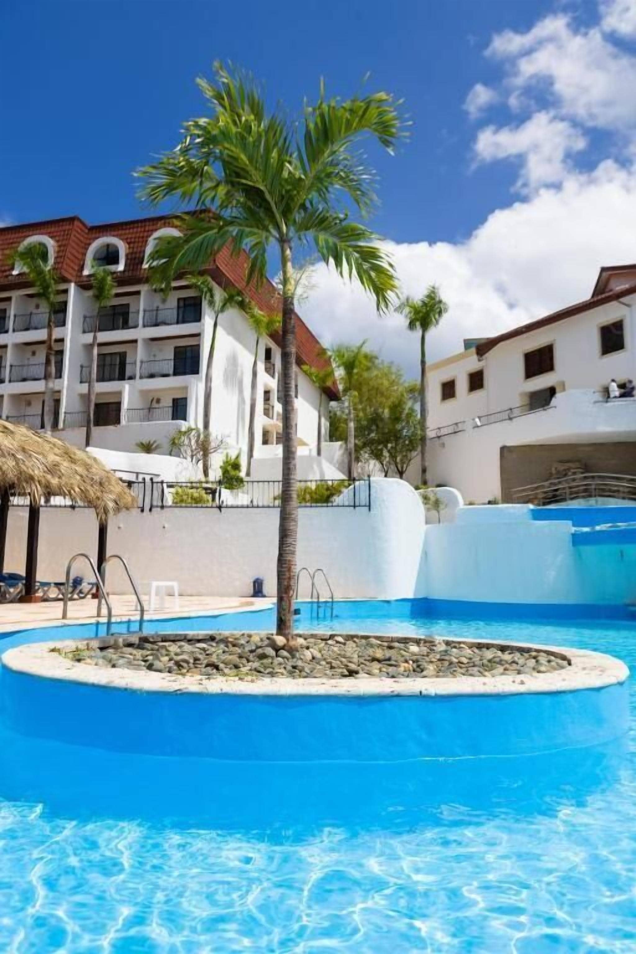 A swimming pool with a palm tree, a building with balconies, and a clear blue sky.