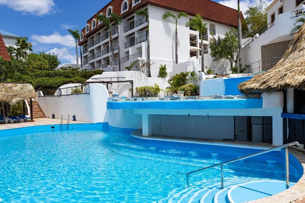 A swimming pool with a blue edge, a thatched roof structure, and a multi-story building with balconies in the background.