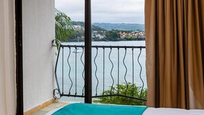 Un balcone con ringhiera in metallo, un letto con copripiumino color acquamarina e vista su un corpo d'acqua e edifici in lontananza.