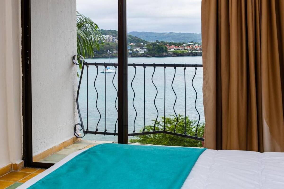 Un balcón con barandilla de metal, una cama con colcha color turquesa, y vista a un cuerpo de agua y edificios lejanos.