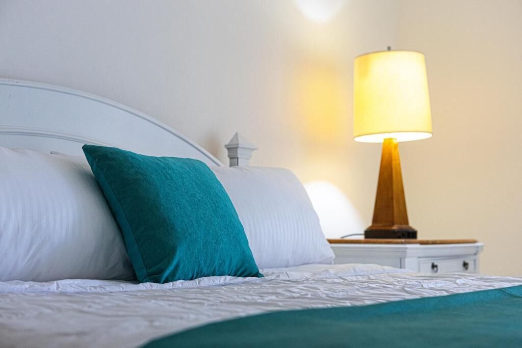 A neatly made bed with white linens and teal pillows, a bedside table with a lamp, and a small white nightstand.