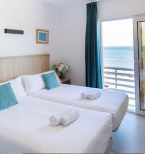 Ein Hotelzimmer mit zwei Betten, Meerblick, einer Schiebetür und einem kleinen Tisch mit Blumen.