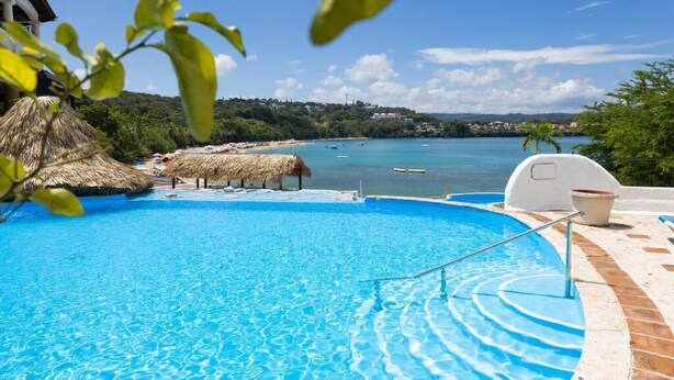 Una piscina con vista sul mare, ombrelloni di paglia e un edificio bianco.