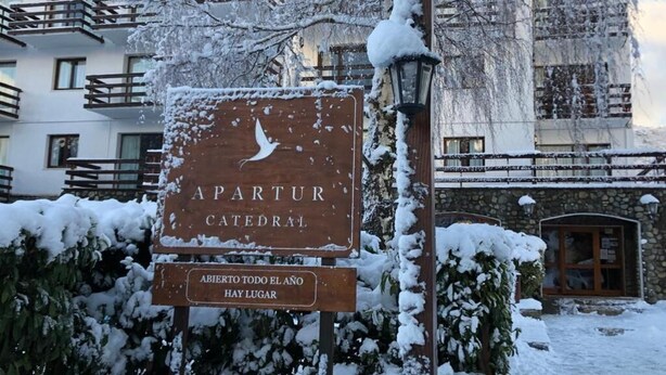 A sign for 'APARTUR CATEDRAL' with a snow-covered environment and a stone building in the background. - Apartur Catedral (San Carlos de Bariloche)