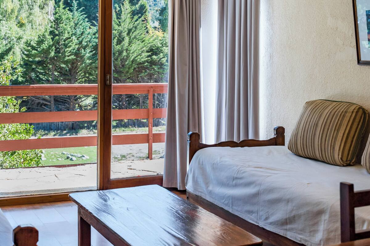 Un dormitorio con cama, un banco de madera, una mesa de madera y vistas a una terraza de madera y árboles a través de un ventanal grande.