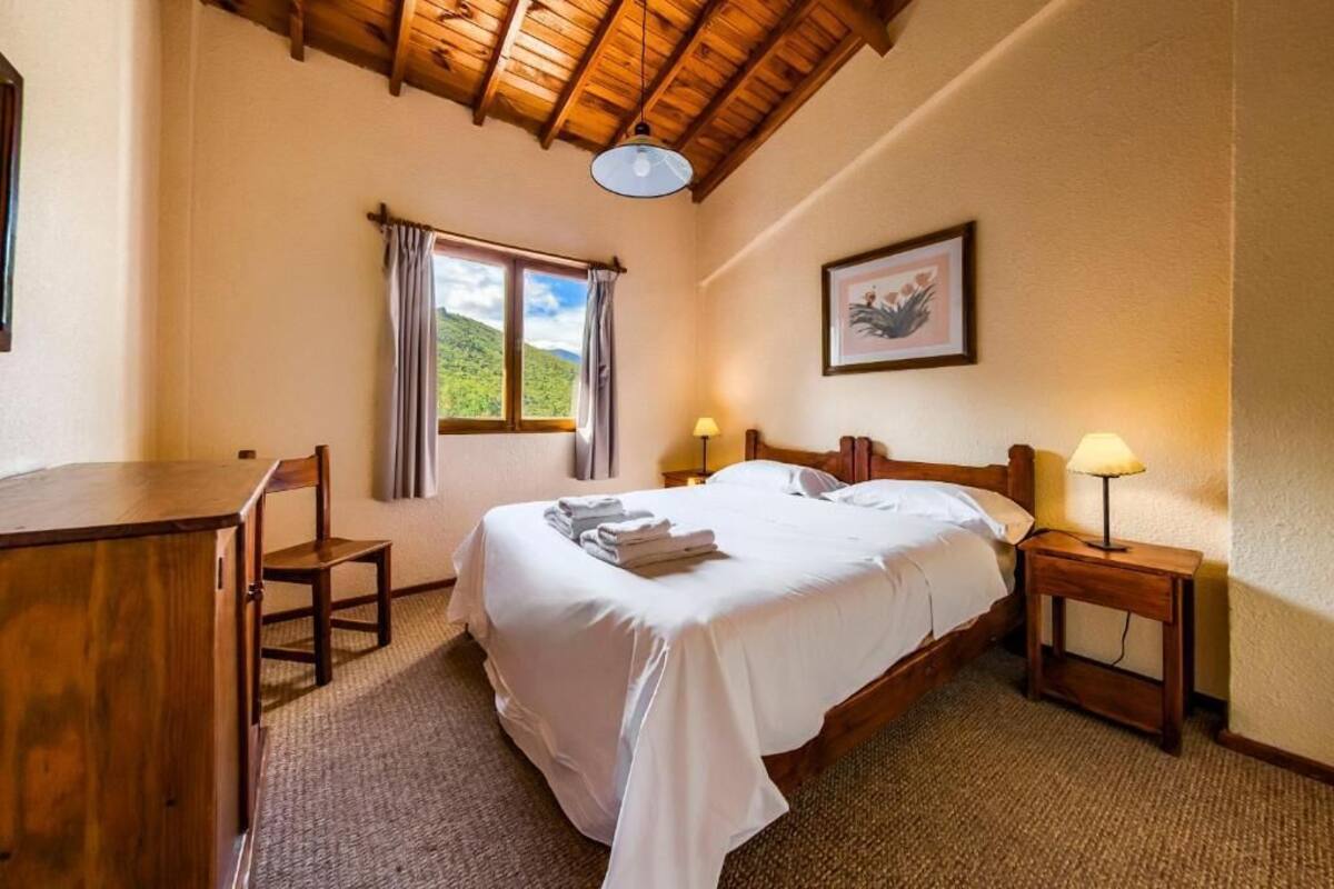 Un dormitorio con una cama de madera, dos mesitas de noche con lámparas, una ventana con cortinas, un cuadro enmarcado en la pared y un escritorio con una silla.
