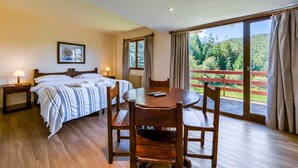 A bedroom with a bed, bedside tables, a round dining table with chairs, a window with curtains, and a view of greenery. - Apartur Catedral (San Carlos de Bariloche)