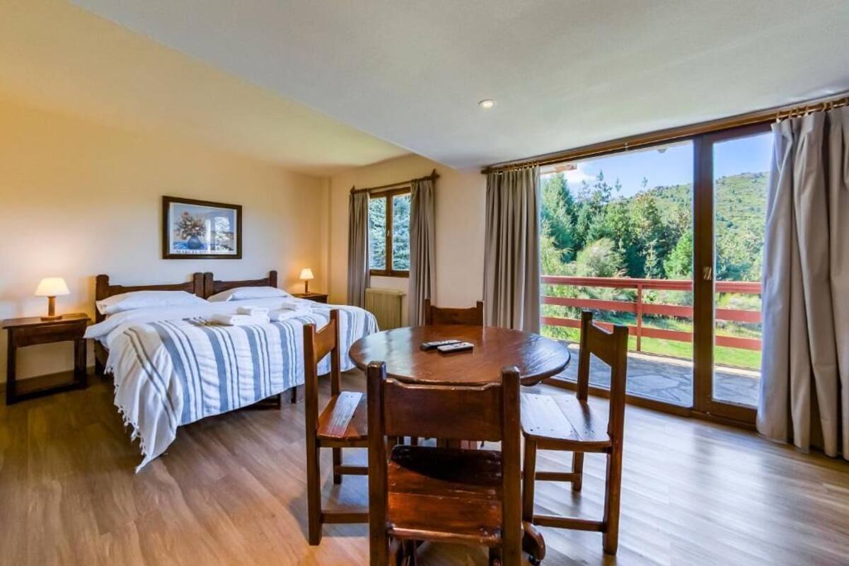 Un dormitorio con cama, mesitas de noche, una mesa redonda con sillas y cortinas en la ventana, y vistas al verdor.