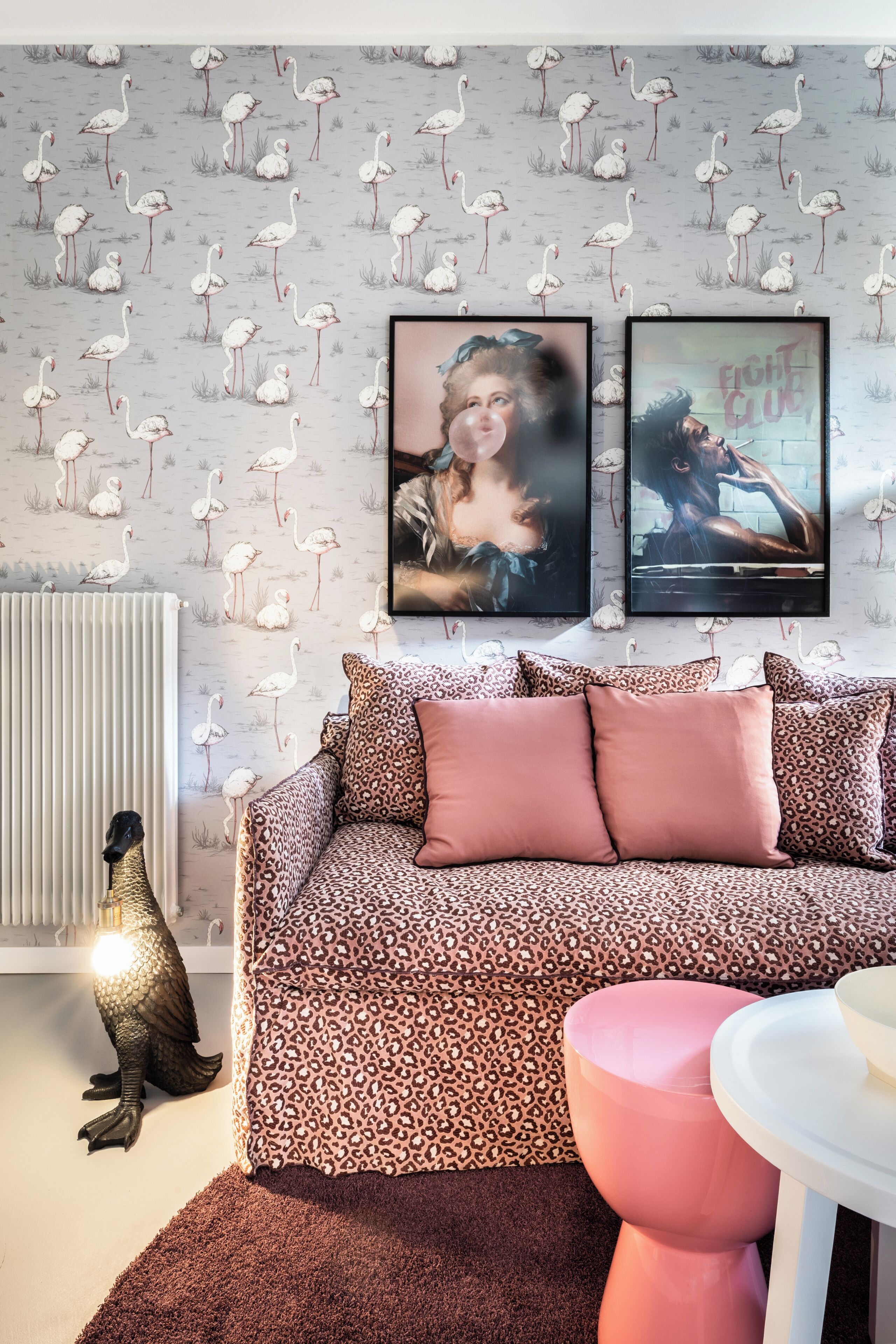 Une pièce avec un papier peint à motifs, un canapé à imprimé léopard, un pouf rose et un radiateur.