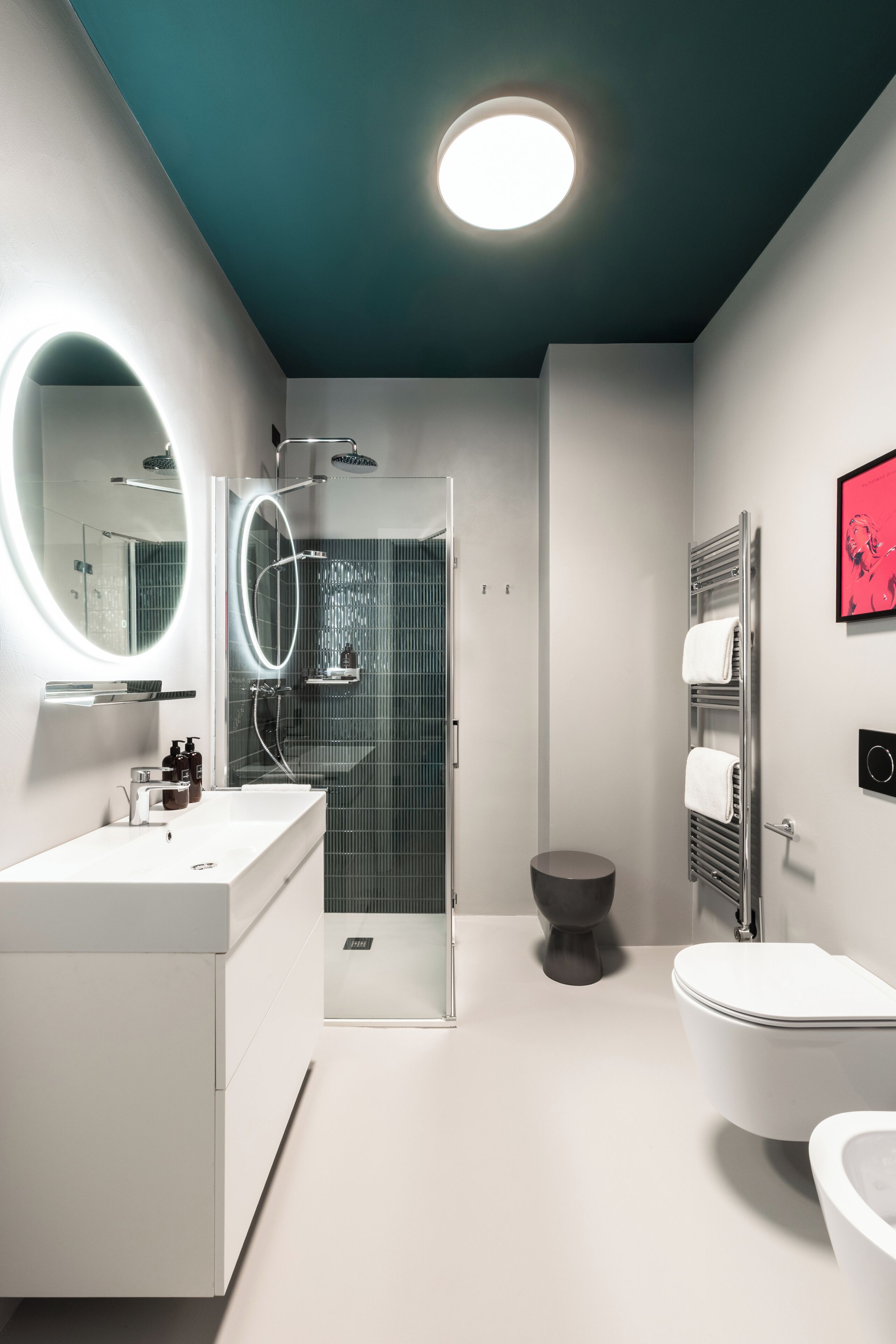 A modern bathroom with a white vanity, a round mirror, a glass shower enclosure, a toilet, and a towel rack.