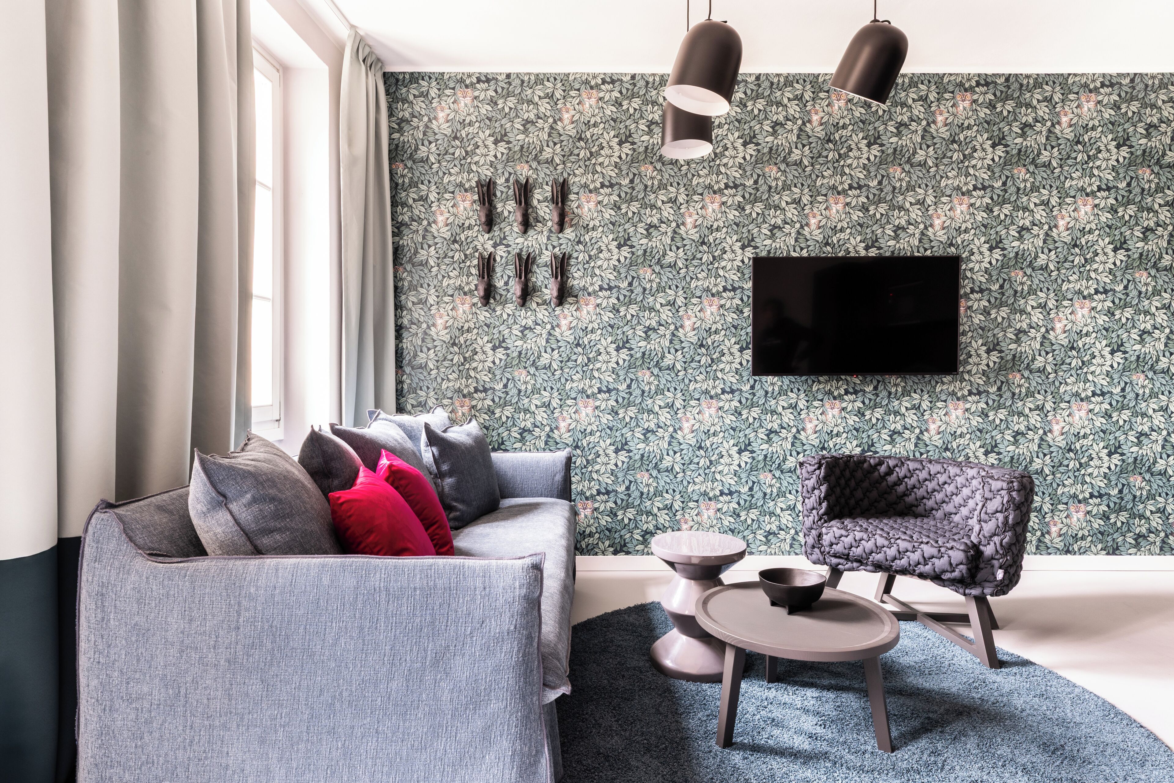 Una sala de estar moderna con un sofá gris, un sillón oscuro, una mesa de centro redonda y un televisor de pantalla plana montado en una pared con estampado floral.