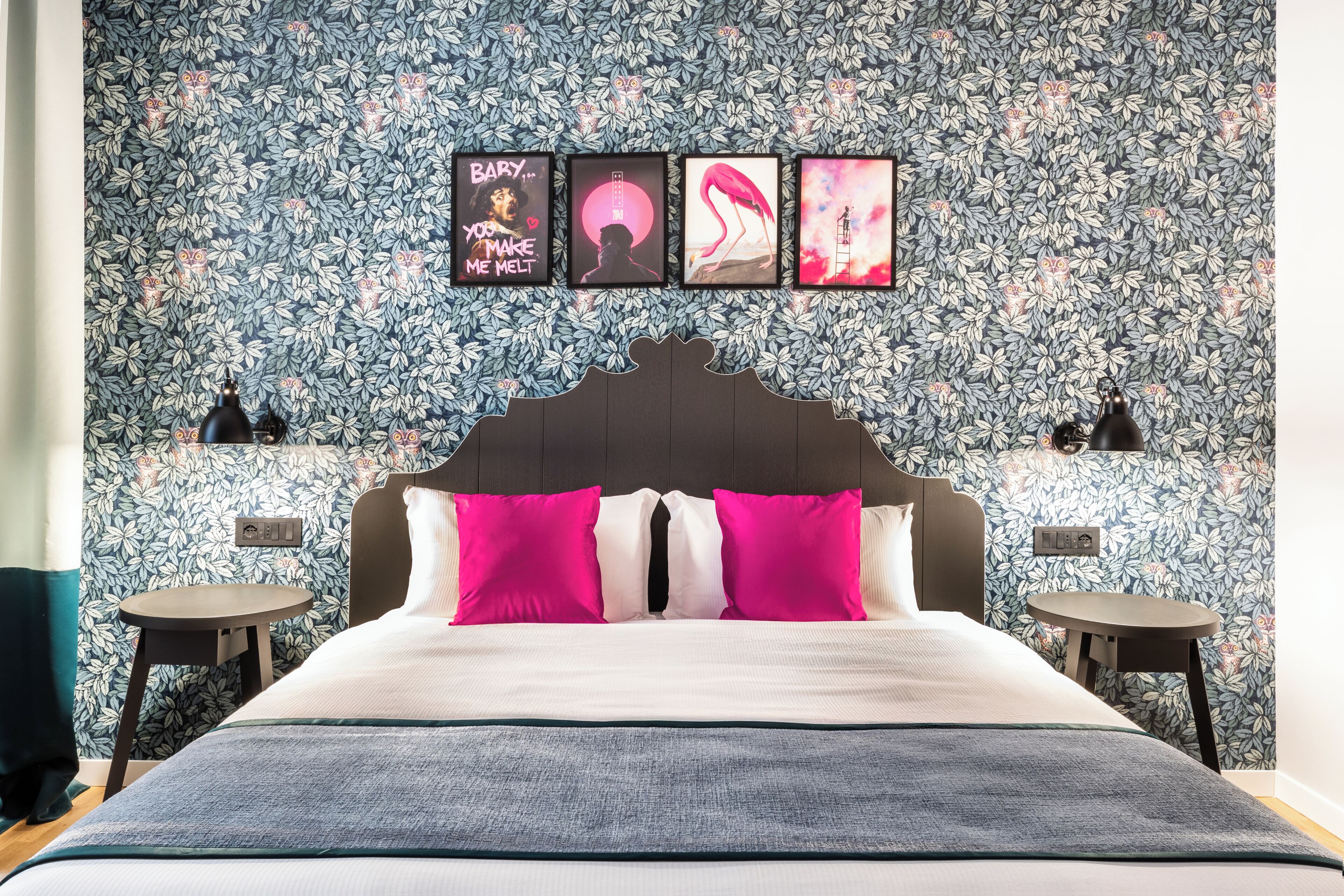 A bedroom with a floral wallpaper, a bed with pink and white pillows, and four framed pictures on the wall.