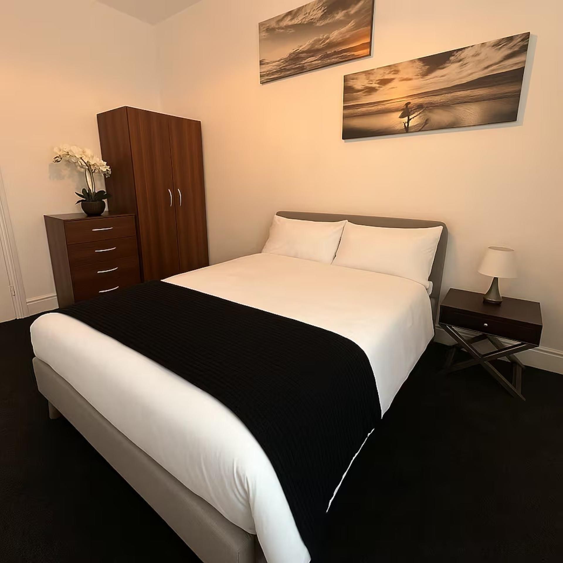 A hotel room with a bed, a nightstand, a lamp, a wardrobe, and two wall-mounted paintings.