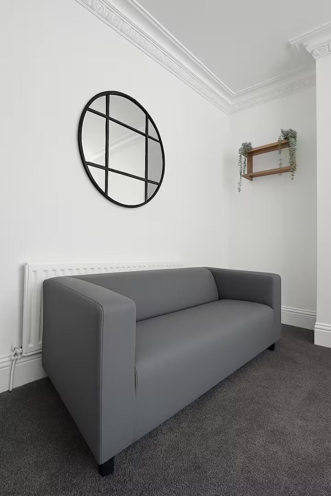 Un sofá gris en una habitación con un radiador blanco, un espejo redondo con marco negro y una pequeña estantería con plantas.