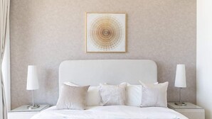 Une chambre à coucher bien rangée, avec un lit, des oreillers, deux tables de chevet avec des lampes et une œuvre d’art accrochée au mur.