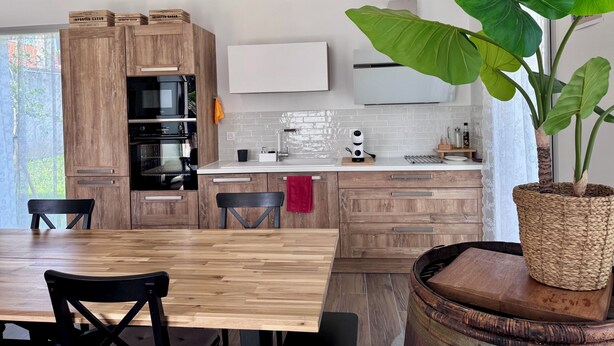Una cocina moderna con armarios de madera, una mesa de comedor, sillas, una planta verde grande y un televisor blanco montado en la pared.