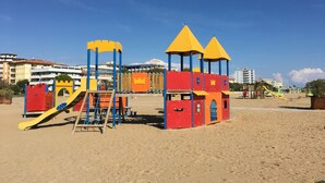 Un parque infantil en la playa con toboganes y estructuras para escalar, con edificios y cielos despejados al fondo.