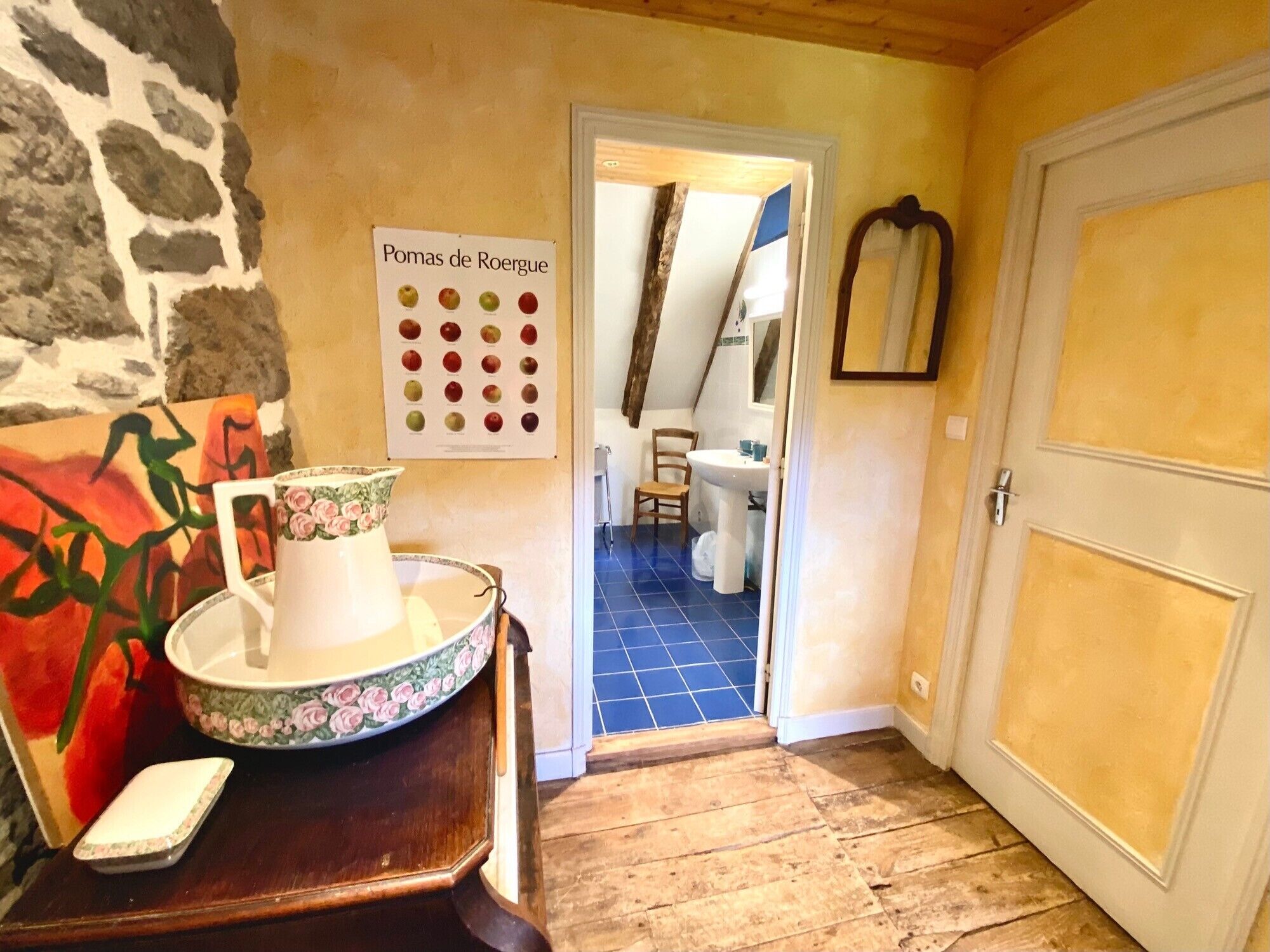 A hallway with a stone wall, a painting, a table with a floral-patterned pitcher, a sign with apple illustrations, and a door leading to a bathroom.
