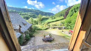 Une vue depuis une fenêtre donnant sur un paysage rural, avec un bâtiment en pierre, une cour verdoyante et une voiture rouge garée à l’extérieur.